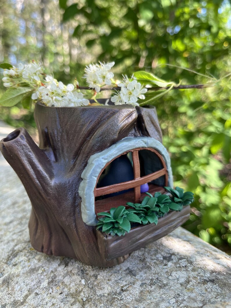 Op de afbeelding: Een grillige, bruine boomstronk-vormige plantenbak met een klein raam en een plantenbak gevuld met groene planten. Witte bloemen en groene bladeren zijn in de plantenbak geplaatst.