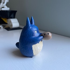 以下が含まれることがあります： 白いお腹と大きな耳を持つ青い漫画のキャラクターのフィギュアで、小さな茶色のオブジェクトを持っています。フィギュアは白い表面にあり、背景はぼやけています。