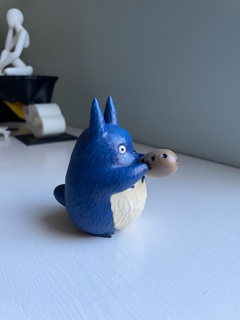 以下が含まれることがあります： 白いお腹と大きな耳を持つ青い漫画のキャラクターのフィギュアで、小さな茶色のオブジェクトを持っています。フィギュアは白い表面にあり、背景はぼやけています。