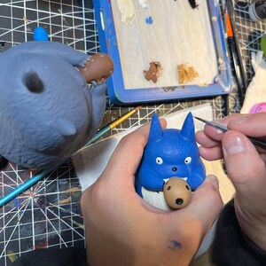 以下が含まれることがあります： 口の中に茶色の物体が入った青い漫画のキャラクターのフィギュアを人がペイントしています。同じキャラクターの2番目の、より大きなフィギュアが背景に見えます。絵の具と青いパレットも画像にあります。
