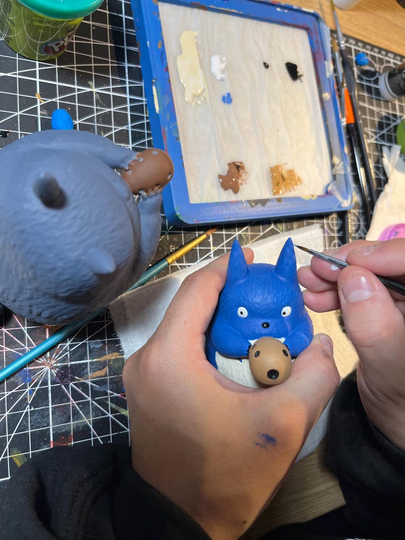 以下が含まれることがあります： 口の中に茶色の物体が入った青い漫画のキャラクターのフィギュアを人がペイントしています。同じキャラクターの2番目の、より大きなフィギュアが背景に見えます。絵の具と青いパレットも画像にあります。