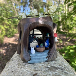 Op de afbeelding: Een gedetailleerde houten sculptuur van een boomstronk met een grillige interieursc&egrave;ne. Binnenin zit een Totoro-figuur met andere personages en miniatuurmeubels. De sculptuur is bruin met een blauw en wit interieur en is versierd met een takje witte bloemen.