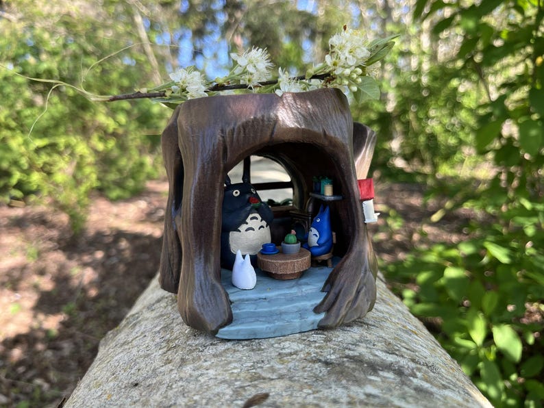 Op de afbeelding: Een gedetailleerde houten sculptuur van een boomstronk met een grillige interieursc&egrave;ne. Binnenin zit een Totoro-figuur met andere personages en miniatuurmeubels. De sculptuur is bruin met een blauw en wit interieur en is versierd met een takje witte bloemen.