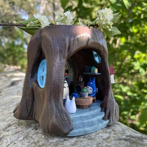 Op de afbeelding: Een grillige miniatuur van een bruine boomstronk met een blauw geschilderd raam en een klein huis. Binnenin zit een Totoro-figuur aan een tafel met cadeaus. Witte bloemen en groen zijn bovenop de stronk geplaatst.