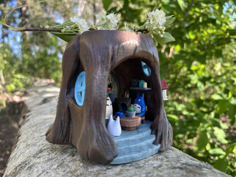 Op de afbeelding: Een grillige miniatuur van een bruine boomstronk met een blauw geschilderd raam en een klein huis. Binnenin zit een Totoro-figuur aan een tafel met cadeaus. Witte bloemen en groen zijn bovenop de stronk geplaatst.