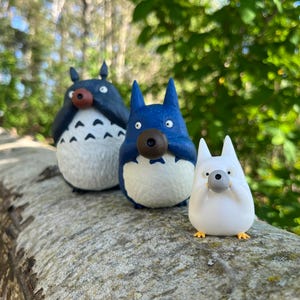 以下が含まれることがあります： 人気アニメのキャラクターの3つの気まぐれなフィギュア。それぞれがユニークな配色です。フィギュアは丸太の上にあり、背景は森です。フィギュアは青、白、濃い青で、鼻は茶色です。