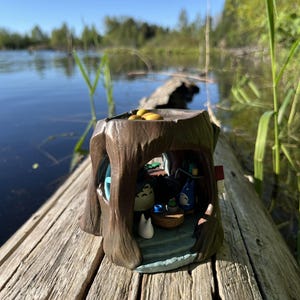 Op de afbeelding: Een gedetailleerd miniatuur boomstronk diorama met personages uit een populaire animatiefilm. De bruine hars sculptuur heeft een blauw interieur met kleine figuren en een tafel. Het diorama staat op een verweerde houten stam.