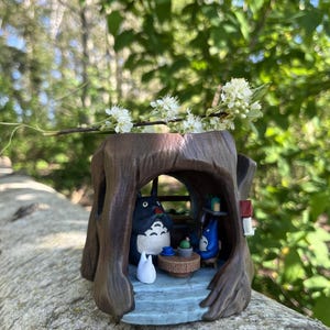 Op de afbeelding: Een grillig, bruin boomstamvormig beeldje met een sc&egrave;ne met Totoro-personages. Binnenin zit een grote Totoro-figuur met een kleine blauwe Totoro en een witte katachtige figuur. Een takje witte bloemen rust bovenop.