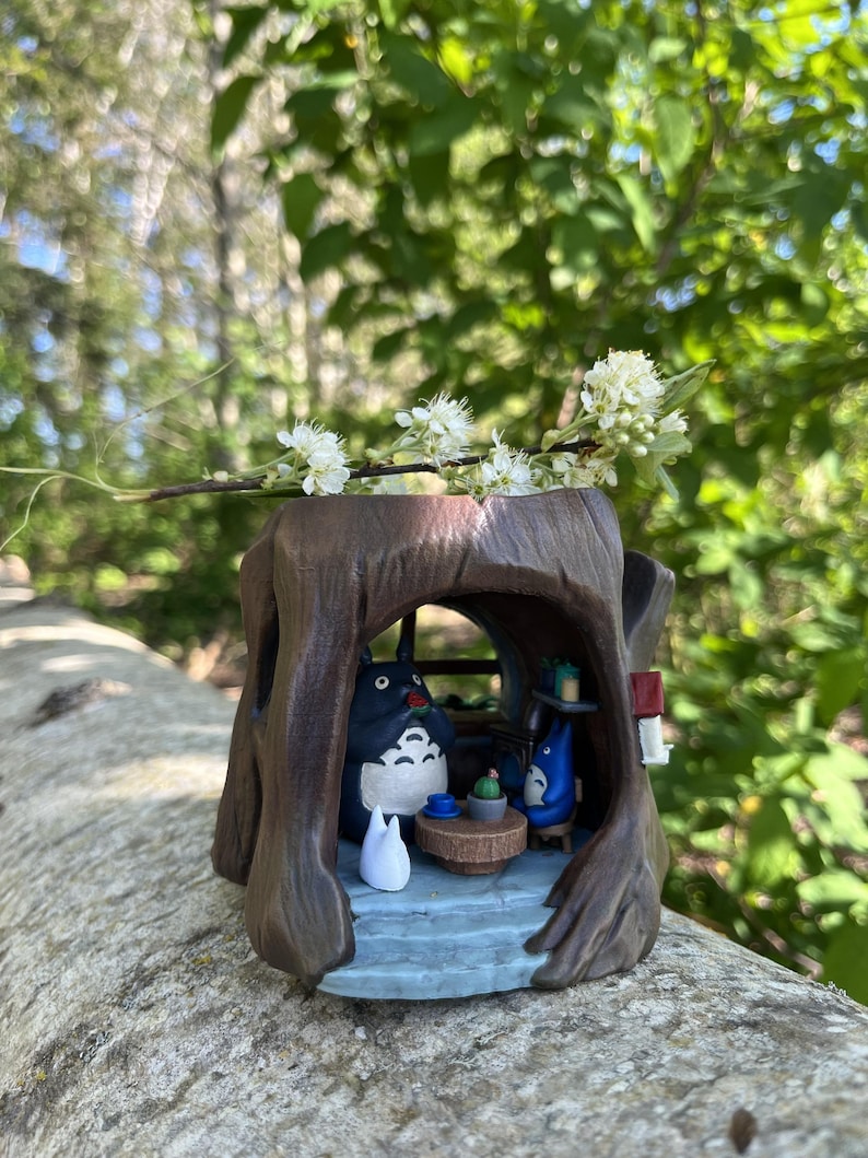 Op de afbeelding: Een grillig, bruin boomstamvormig beeldje met een sc&egrave;ne met Totoro-personages. Binnenin zit een grote Totoro-figuur met een kleine blauwe Totoro en een witte katachtige figuur. Een takje witte bloemen rust bovenop.