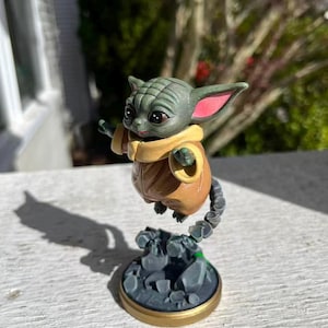 Op de afbeelding: Een beeldje van een groen alien-personage met grote oren en grote ogen, gekleed in een bruine jas en een gele sjaal. Het beeldje is bevestigd aan een grijze basis met een gouden rand en een rotsachtige textuur.