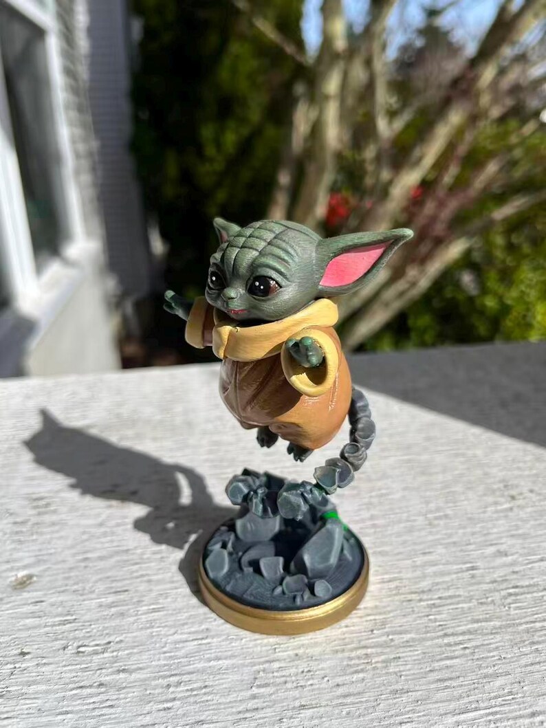 Op de afbeelding: Een beeldje van een groen alien-personage met grote oren en grote ogen, gekleed in een bruine jas en een gele sjaal. Het beeldje is bevestigd aan een grijze basis met een gouden rand en een rotsachtige textuur.