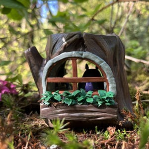 Op de afbeelding: Een grillig miniatuur boomstronk huis met een raam, een groene plantenbak en een klein figuur binnenin. Het huis is bruin met een lichtgroene rand rond het raam. De sc&egrave;ne speelt zich buiten af met een vage groene achtergrond.