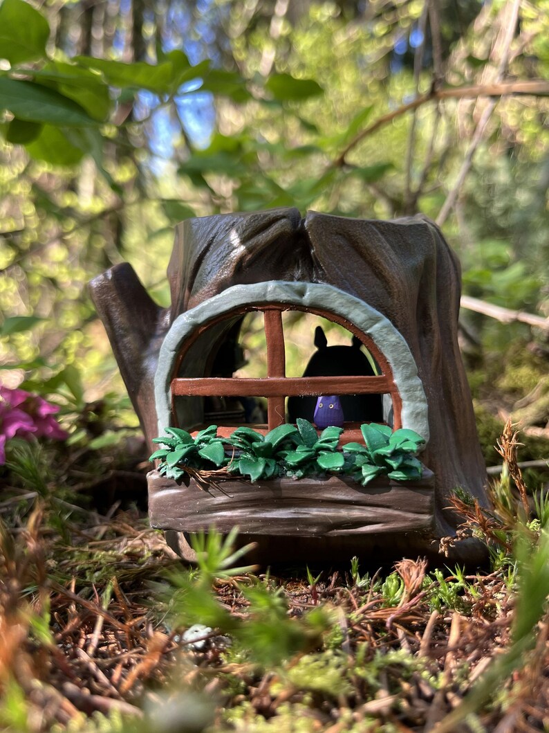 Op de afbeelding: Een grillig miniatuur boomstronk huis met een raam, een groene plantenbak en een klein figuur binnenin. Het huis is bruin met een lichtgroene rand rond het raam. De sc&egrave;ne speelt zich buiten af met een vage groene achtergrond.