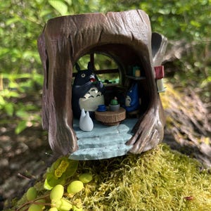 Op de afbeelding: Gedetailleerd beeldje dat een sc&egrave;ne uit de animatiefilm, Mijn buurman Totoro, uitbeeldt. De bruine boomstamvormige structuur herbergt Totoro en andere personages. De sc&egrave;ne omvat een tafel, een raam en een klein wit wezen. Het beeldje staat op een bed van groen mos.
