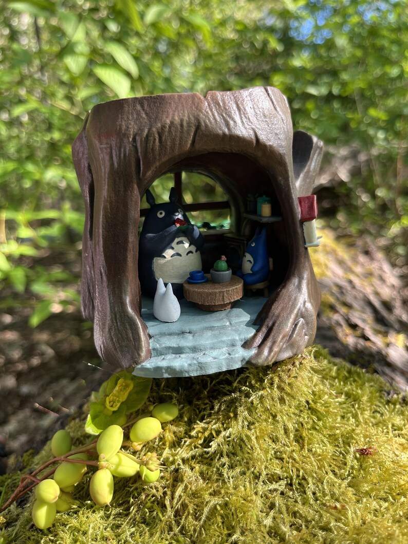 Op de afbeelding: Gedetailleerd beeldje dat een sc&egrave;ne uit de animatiefilm, Mijn buurman Totoro, uitbeeldt. De bruine boomstamvormige structuur herbergt Totoro en andere personages. De sc&egrave;ne omvat een tafel, een raam en een klein wit wezen. Het beeldje staat op een bed van groen mos.