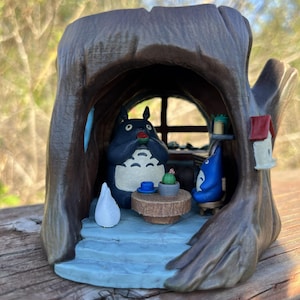 Op de afbeelding: Een grillig beeldje dat een sc&egrave;ne in een boomstam uitbeeldt. Het interieur heeft een tafel met een cactus en twee Totoro-figuren. De boomstam is bruin, met een blauw interieur en een klein vogelhuisje.