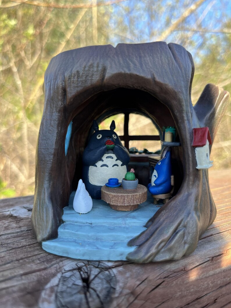 Op de afbeelding: Een grillig beeldje dat een sc&egrave;ne in een boomstam uitbeeldt. Het interieur heeft een tafel met een cactus en twee Totoro-figuren. De boomstam is bruin, met een blauw interieur en een klein vogelhuisje.