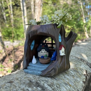 Op de afbeelding: Een grillige, bruine boomstamplanter met een Totoro-figuur binnenin. De planter heeft een klein vogelhuisje en blauwe accenten. Witte bloemen zijn bovenop gerangschikt en cre&euml;ren een charmant, door de natuur ge&iuml;nspireerd decorstuk.