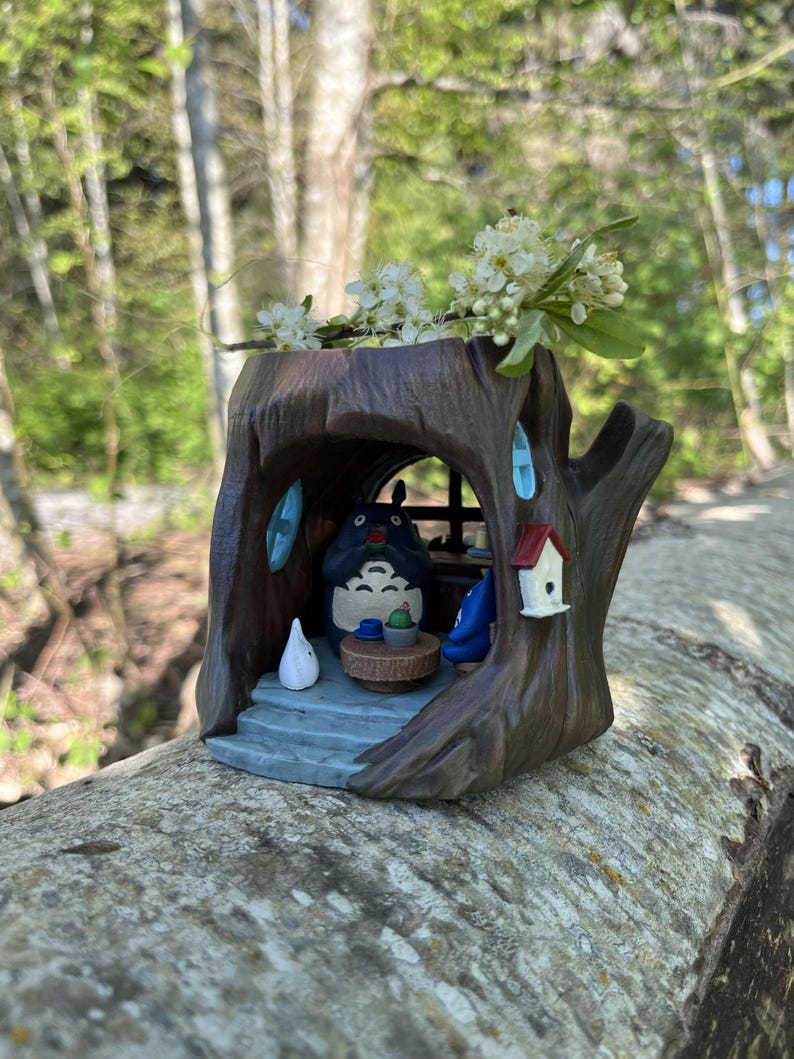 Op de afbeelding: Een grillige, bruine boomstamplanter met een Totoro-figuur binnenin. De planter heeft een klein vogelhuisje en blauwe accenten. Witte bloemen zijn bovenop gerangschikt en cre&euml;ren een charmant, door de natuur ge&iuml;nspireerd decorstuk.