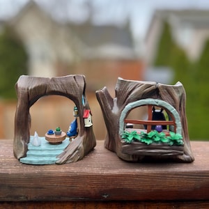 Op de afbeelding: Twee grillige boomstronk-plantenbakken. E&eacute;n heeft een miniatuursc&egrave;ne met een tafel en stoelen, de andere heeft een raam met een kat erin. Beide zijn geschilderd in bruin-, groen- en blauwtinten, wat een charmante, sprookjesachtige esthetiek cre&euml;ert.