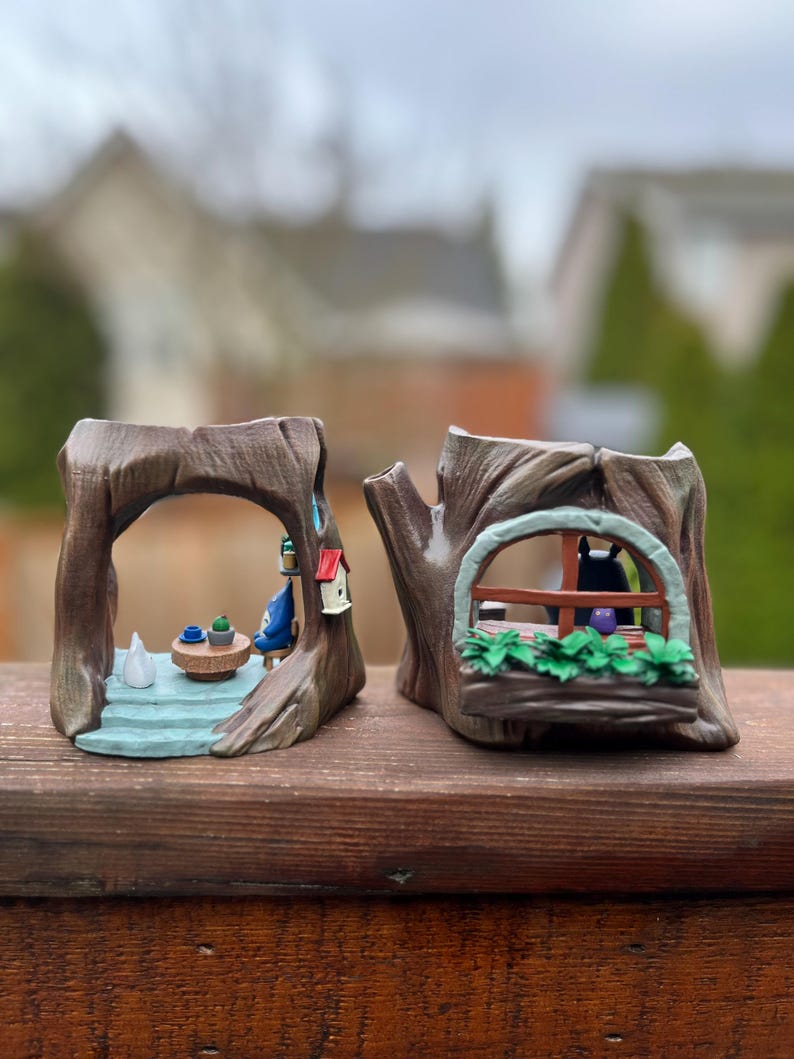 Op de afbeelding: Twee grillige boomstronk-plantenbakken. E&eacute;n heeft een miniatuursc&egrave;ne met een tafel en stoelen, de andere heeft een raam met een kat erin. Beide zijn geschilderd in bruin-, groen- en blauwtinten, wat een charmante, sprookjesachtige esthetiek cre&euml;ert.