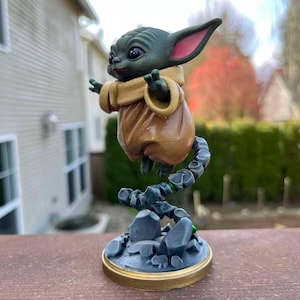 Op de afbeelding: Een groene figuur van een personage met grote oren en een bruine jurk, zwevend boven een rotsachtige basis. De figuur heeft grote ogen en poseert met uitgestrekte armen. De basis is grijs met een gouden rand en de figuur wordt ondersteund door een grijze, rotsachtige structuur.