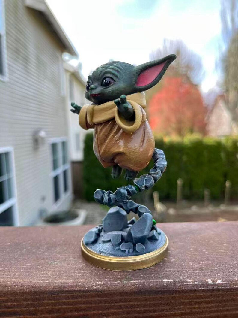 Op de afbeelding: Een groene figuur van een personage met grote oren en een bruine jurk, zwevend boven een rotsachtige basis. De figuur heeft grote ogen en poseert met uitgestrekte armen. De basis is grijs met een gouden rand en de figuur wordt ondersteund door een grijze, rotsachtige structuur.