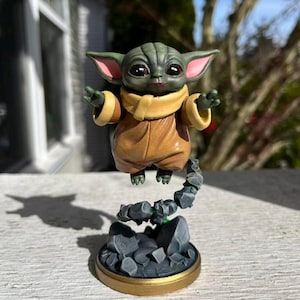 Op de afbeelding: Gedetailleerd beeldje van een groenhuidige alien met grote ogen en puntige oren, gekleed in een bruine jurk en een beige sjaal. De figuur zweeft boven een rotsachtige basis met een goudkleurige rand, waardoor een zwevend effect ontstaat.