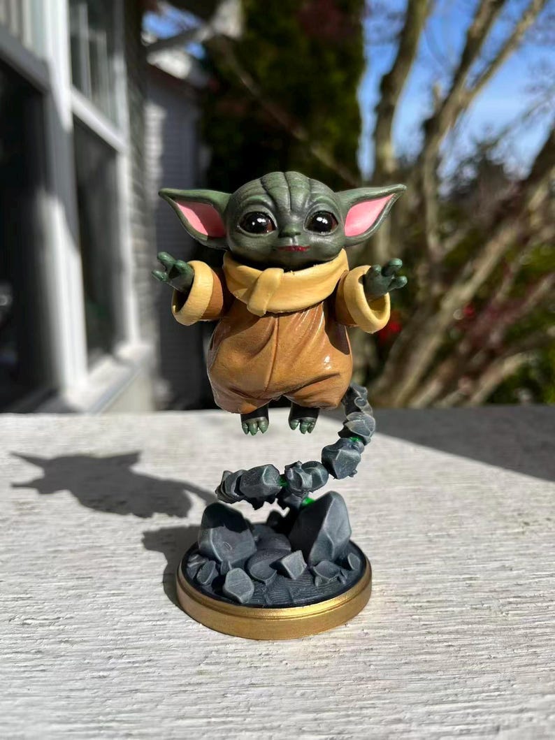 Op de afbeelding: Gedetailleerd beeldje van een groenhuidige alien met grote ogen en puntige oren, gekleed in een bruine jurk en een beige sjaal. De figuur zweeft boven een rotsachtige basis met een goudkleurige rand, waardoor een zwevend effect ontstaat.