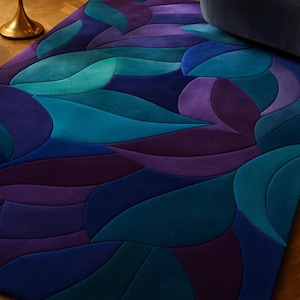 Puede incluir: Una alfombra grande y abstracta con un patrón de hojas en tonos turquesa, azul y morado. La alfombra está colocada sobre un suelo de madera, con una pequeña mesa auxiliar dorada y un sofá azul oscuro en el fondo.