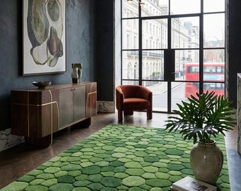 Op de afbeelding: Een moderne woonkamer met een groen, getextureerd tapijt met een geometrisch patroon. Een houten kast met gouden accenten en een roestkleurige fauteuil zijn ook aanwezig. Een groot raam biedt uitzicht op een stadsstraat.