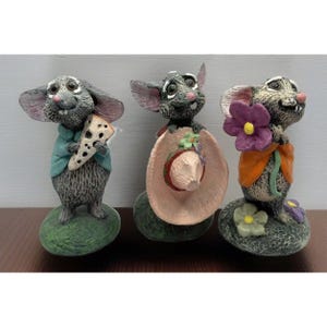 May include: Three whimsical mouse figurines. One mouse holds a slice of cheese, another a pink hat, and the third a purple flower. Each mouse is on a green base, with detailed features and colorful clothing.