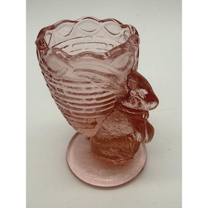 Peut inclure: Un vase décoratif en verre rose avec un motif de lapin. Le vase a une base ronde et un extérieur texturé et détaillé. Le haut du vase a un bord festonné. Le vase est en verre rose.