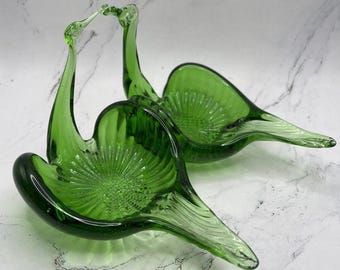 May include: Two green glass swan-shaped decorative bowls. The bowls have a textured, fan-like design in the center and a long, curved neck. The bowls are made of a deep green glass and are likely vintage.