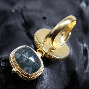 May include: A gold-colored ring with a large, faceted, oval-shaped green stone. The ring has a decorative gold band and a smaller, matching stone. The ring is set against a dark background.