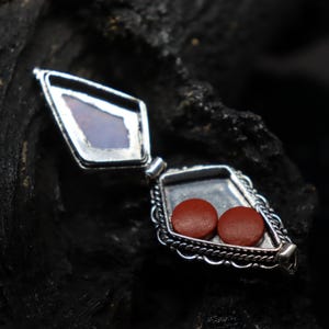 Pu&ograve; includere: Ciondolo aperto color argento a forma di diamante, che rivela due piccoli oggetti rotondi marrone-rossastri. Il ciondolo ha un bordo decorativo a forma di corda e un coperchio incernierato. Lo sfondo &egrave; una superficie scura e strutturata.
