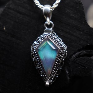 Pu&ograve; includere: Una collana con pendente in argento con una pietra preziosa iridescente a forma di diamante. La pietra preziosa mostra sfumature di blu, verde e viola. Il pendente &egrave; incastonato in una montatura d'argento ornata e dettagliata.