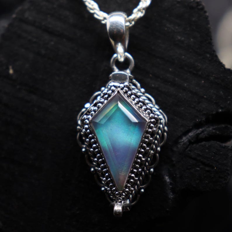 Pu&ograve; includere: Una collana con pendente in argento con una pietra preziosa iridescente a forma di diamante. La pietra preziosa mostra sfumature di blu, verde e viola. Il pendente &egrave; incastonato in una montatura d'argento ornata e dettagliata.