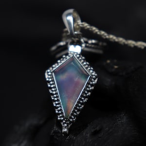 Pu&ograve; includere: Un ciondolo d'argento con una gemma iridescente a forma di diamante. La gemma mostra colori come viola, blu e verde. Il ciondolo &egrave; attaccato a una catena d'argento e ha un bordo dettagliato con perline.