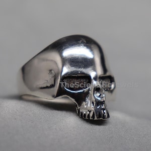 May include: A polished silver-toned ring shaped like a human skull. The skull features dark, recessed eye sockets and a detailed nasal cavity and teeth. The text TheScientificJewels is visible across the eye sockets of the skull ring.