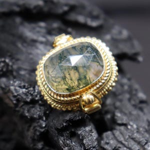 May include: A gold-colored pendant with a large, faceted, moss agate stone. The stone is a mix of clear and green, with visible inclusions. The pendant has a decorative rope-like border and small loops for a chain.