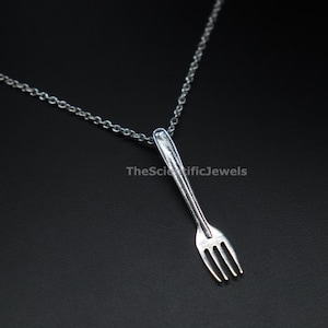 May include: A silver-toned metal necklace featuring a miniature fork pendant suspended from a delicate chain. The pendant is vertically oriented and includes the text TheScientificJewels printed in white across the center of the fork handle.