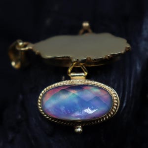 May include: A gold-colored pendant with an oval, faceted, iridescent gemstone. The gemstone is framed by a twisted rope design. The pendant has a unique, irregular top piece and a small loop for a chain.