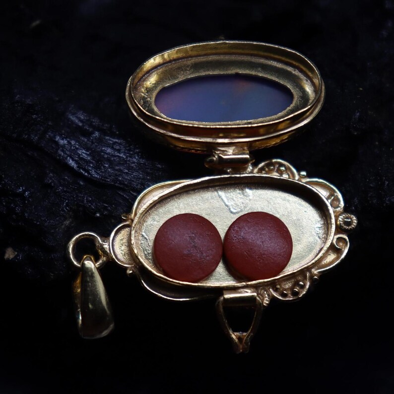 May include: A gold-colored, ornate pendant with an oval-shaped compartment containing two small, round, reddish-brown objects. The pendant has a hinged top with a translucent, oval-shaped stone. The background is dark.
