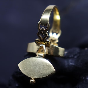 May include: A gold-colored ring with an ornate design. The ring features a decorative element with a teardrop shape and a small, round detail at the bottom. The band has an intricate pattern, and the ring is set against a dark background.