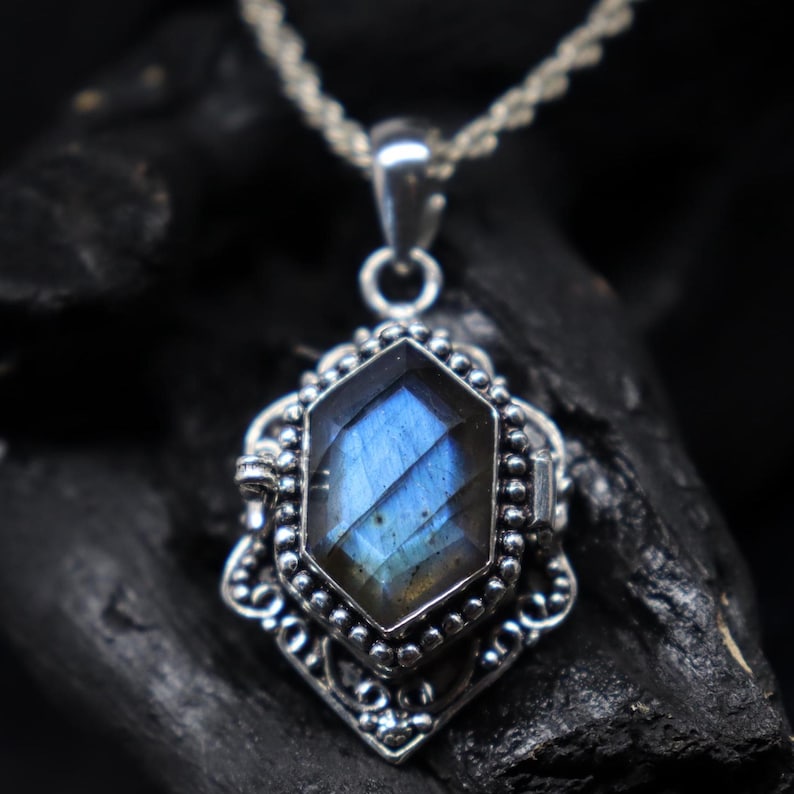 May include: A silver pendant necklace featuring a faceted, blue gemstone. The gemstone is set in an ornate, filigree-style frame with a beaded border. The necklace has a delicate, twisted chain.