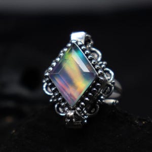 Puede incluir: Un anillo de plata con una gran piedra preciosa iridiscente en forma de diamante. La piedra preciosa muestra un espectro de colores, incluyendo verde, amarillo y morado. El anillo tiene un engaste ornamentado y detallado con un borde de cuentas y adornos decorativos.