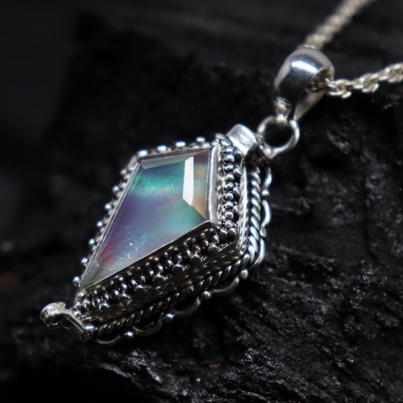 Pu&ograve; includere: Una collana con pendente in argento con una gemma geometrica e iridescente. Il pendente ha una forma a diamante con una montatura in argento dettagliata e ornata. La gemma mostra una gamma di colori, tra cui verde, viola e blu. Anche la catena della collana &egrave; in argento.
