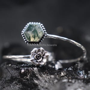May include: A silver open cuff bracelet featuring a hexagonal moss agate stone in a beaded setting on one end and a sculpted metal rose on the other. The band has a hammered texture. The text TheScientificJewels is visible on the stone.