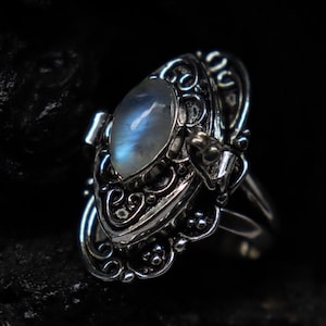 Könnte beinhalten: Ein silberner Ring mit einem ovalen, irisierenden Mondstein in der Mitte. Der Ring hat verzierte Filigranarbeiten und dekorative Akzente. Der Mondstein hat einen blauen und weißen Schimmer. Der Ring ist vor einem dunklen Hintergrund platziert.